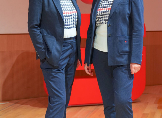 Zwei Mitarbeiterinnen einer Sparkassen-Filiale in dunkelblauen Business-Outfits mit gemusterten Shirts und weißen Sneakern